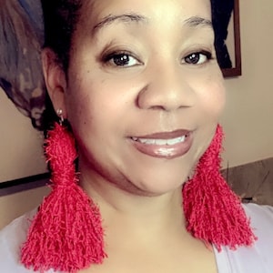 May include: A woman wearing a light purple v-neck top with long red tassel earrings.