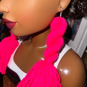 May include: A pair of pink yarn tassel earrings with a braided design. The earrings are hanging from silver hooks.
