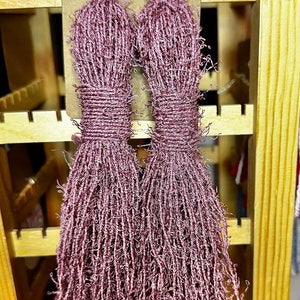 May include: A pair of long, pink tassel earrings with a metallic sheen. The earrings are displayed on a wooden stand.