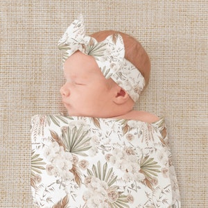 May include: A baby swaddled in a white swaddle with a floral pattern. The swaddle has a white background with brown, green, and beige floral designs. The baby is wearing a white headband with a floral pattern. The headband has a white background with brown, green, and beige floral designs.