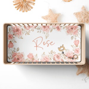 May include: A white crib sheet with a pink floral pattern and the name "Rose" printed in the center. The sheet is in a crib with wooden slats.