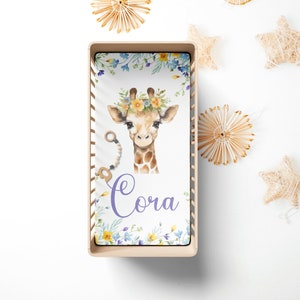 May include: A white crib sheet with a watercolor illustration of a giraffe wearing a flower crown. The sheet has a purple floral border and the name "Cora" written in purple script.
