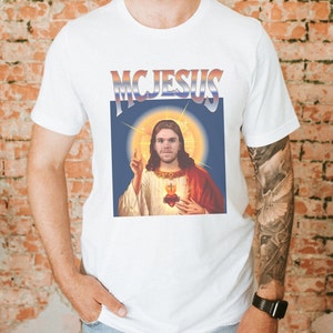 May include: White t-shirt with a graphic of a man with long hair and a beard, wearing a red robe and holding a heart. The graphic is in a retro style with a blue background and the text "MCJESUS" above it.