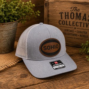 May include: A light gray trucker hat with a white mesh back and a brown oval leather patch on the front featuring the text SOHIO in black. The hat includes a black and white sticker on the brim with the text Richardson 112 Trucker Adjustable.