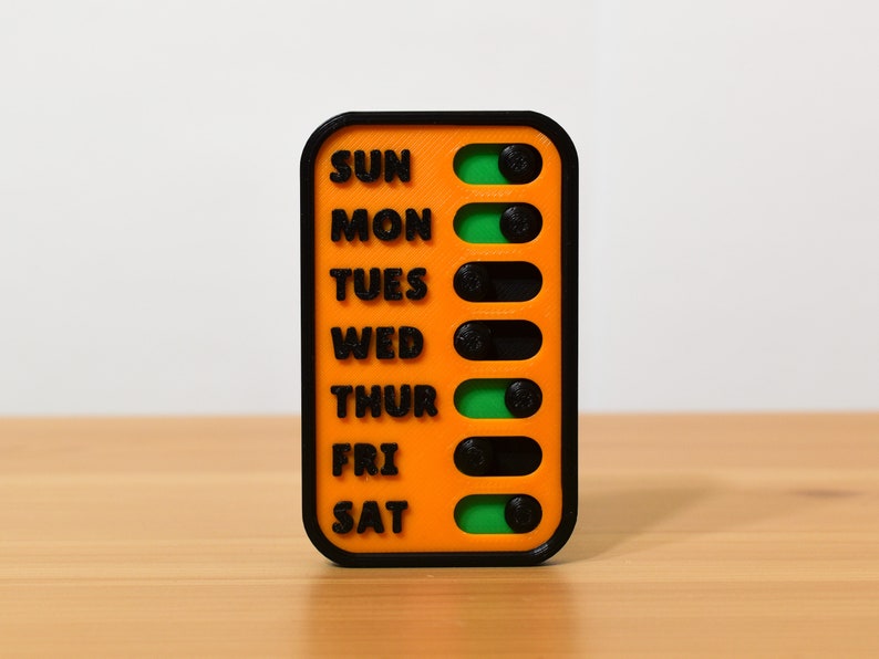Close up view of the magnetic checklist with seven sliders, each labeled with a day of the week, indicating pending or complete status.