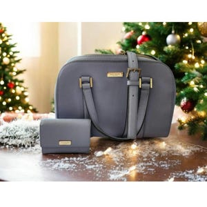 Könnte beinhalten: Eine graue Handtasche und eine passende Geldbörse mit goldfarbener Hardware. Die Handtasche hat einen oberen Griff und einen Schulterriemen. Die Geldbörse ist klein und rechteckig. Beide Artikel tragen den Markennamen. Auf einer Holzoberfläche mit Weihnachtsbaum im Hintergrund.