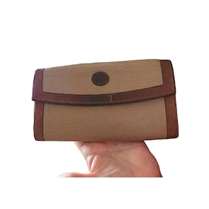 May include: A tan and brown leather clutch bag with a flap closure. The bag has a textured tan body, brown leather trim, and a circular brown accent. The clutch is held in a hand.