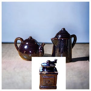 May include: A set of three ceramic kitchen items: a small teapot, a creamer, and a coffee grinder. The teapot and creamer are dark brown with a glossy finish. The coffee grinder is a dark brown color with a wooden-like appearance.