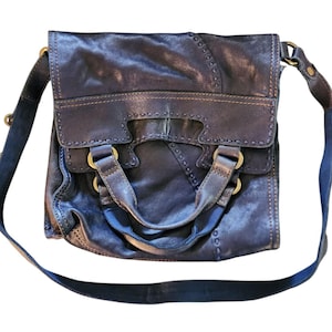 Puede incluir: Bolso bandolera de cuero azul oscuro con cierre de solapa y correa larga ajustable. El bolso presenta detalles de costura, herrajes color latón y un asa superior. El cuero parece desgastado, dándole un aspecto vintage.