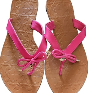 May include: A pair of pink flip-flops with a bow detail. The sandals have a brown sole with a textured pattern. The straps are a vibrant pink color, and the bow has gold-colored accents. The flip-flops are designed for casual wear.