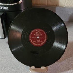 May include: A black vinyl record with a red and white label. The label reads "Victor" and "FAUST: WALTZES" by the Boston "Pops" Orchestra. The record is displayed on a silver stand.