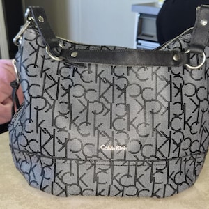 May include: A black and gray Calvin Klein shoulder bag with a repeating CK logo pattern. The bag has a black strap and silver hardware. The bag is sitting on a light-colored surface.