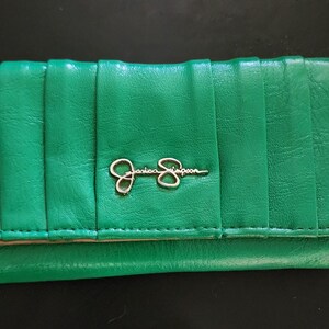 May include: A bright green wallet with a vertical ribbed design. The wallet has a gold-colored "Jessica Simpson" logo on the front. The wallet is closed and appears to be made of leather.