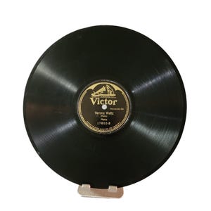 May include: A black vinyl record with a gold and black label that reads "Victor" and "Verona Waltz". The record is held by a silver metal stand. The record is a vintage music item.