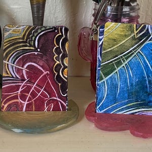 Pode incluir: Quatro peças de arte abstrata, cada uma montada em uma base decorativa. A arte apresenta padrões em espiral em tons de amarelo, roxo, vermelho, azul e verde. As bases são em cores complementares, criando uma exibição coesa. As obras de arte são exibidas em uma prateleira.