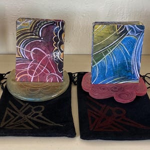 Pode incluir: Quatro porta-cartões decorativos com designs de arte abstrata. Cada suporte tem um esquema de cores único, incluindo roxo, verde-azulado e vermelho, e fica em cima de uma bolsa de veludo preta com um design geométrico. Os cartões apresentam padrões coloridos e rodopiantes.