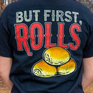 Puede incluir: Una camiseta negra con el texto "But First, Rolls" en letras blancas y rojas. La camiseta presenta un gráfico de tres rollos de color marrón dorado.