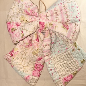May include: A large decorative bow made from quilted fabric featuring a patchwork design. The textile displays various patterns including pink roses, light blue sections, and a grid print. A small jute twine loop is attached to the center knot.