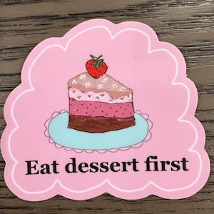 May include: A pink sticker with a scalloped edge features a slice of cake topped with a strawberry. The cake sits on a blue plate, and the text "Eat dessert first" is below the cake.