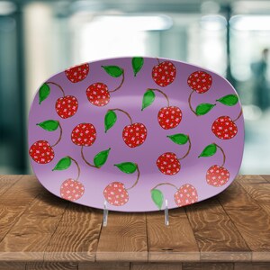 May include: A decorative, oval-shaped plate with a lavender background. The plate is adorned with a repeating pattern of red cherries with white polka dots, green leaves, and brown stems. The plate is displayed on a wooden surface.
