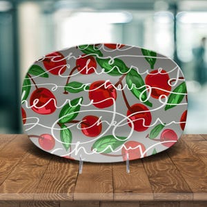May include: Oval ceramic serving platter with a gray background, featuring a pattern of red cherries, green leaves, and white cursive text that reads "cherry". The platter is displayed on a wooden surface.