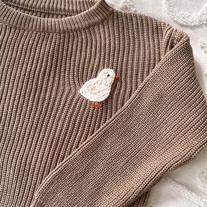 May include: A brown knit sweater with a ribbed texture. A white, embroidered chick with an orange beak and feet is sewn onto the sweater. The sweater has a crew neck and long sleeves.