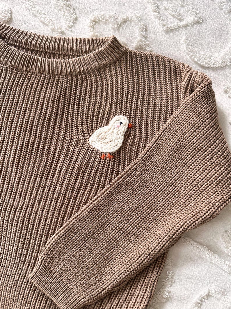 May include: A brown knit sweater with a ribbed texture. A white, embroidered chick with an orange beak and feet is sewn onto the sweater. The sweater has a crew neck and long sleeves.