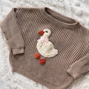 May include: Brown knit sweater with a crew neck and long sleeves. The sweater features a white goose applique with an orange beak and feet, and a pink bow. The sweater is made of a ribbed knit material.