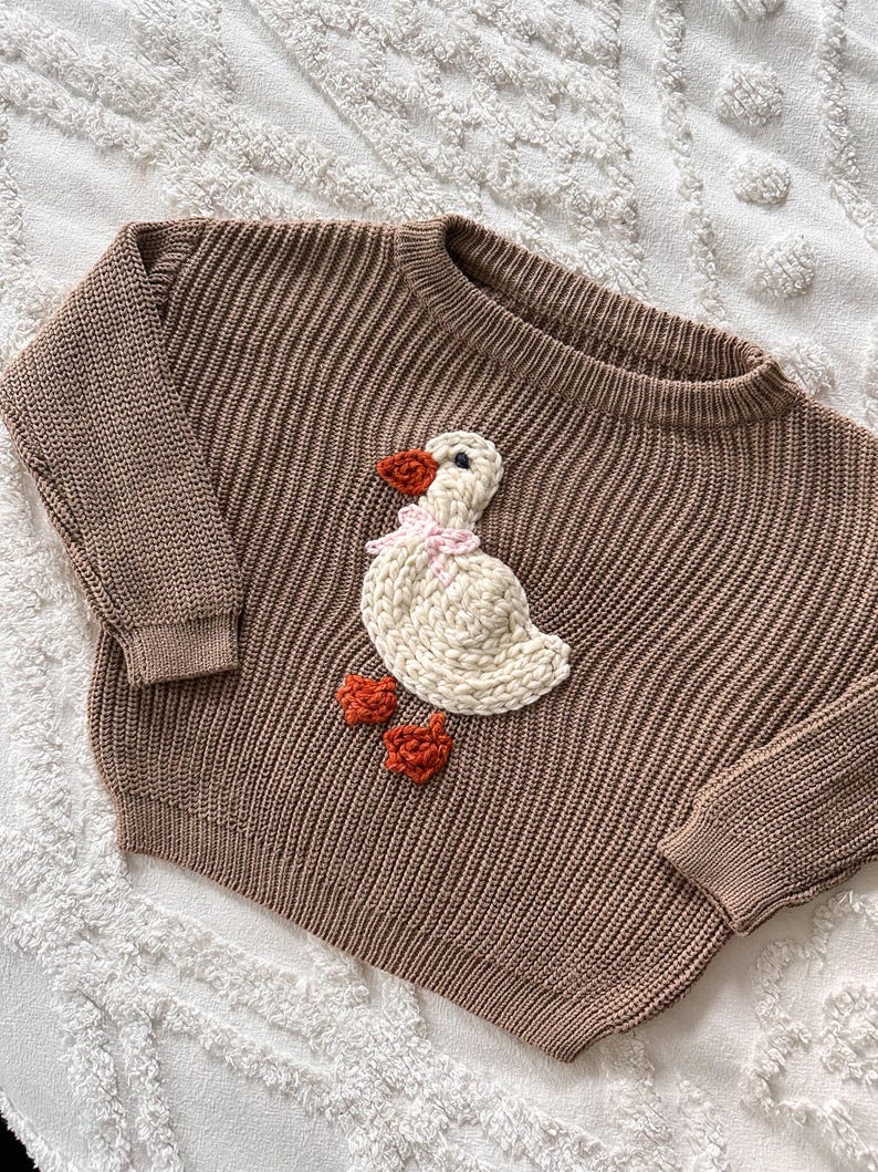 May include: Brown knit sweater with a crew neck and long sleeves. The sweater features a white goose applique with an orange beak and feet, and a pink bow. The sweater is made of a ribbed knit material.
