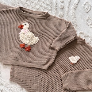 May include: Two brown knit sweaters with a ribbed texture. One sweater features a white duck applique with a pink bow and orange feet. The second sweater has a smaller duck applique. The sweaters are displayed on a white textured surface.