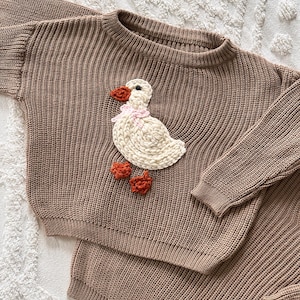 May include: A brown knit sweater and shorts set. The sweater features a white duck applique with an orange beak and feet, and a pink bow. The set is made of a ribbed knit fabric.