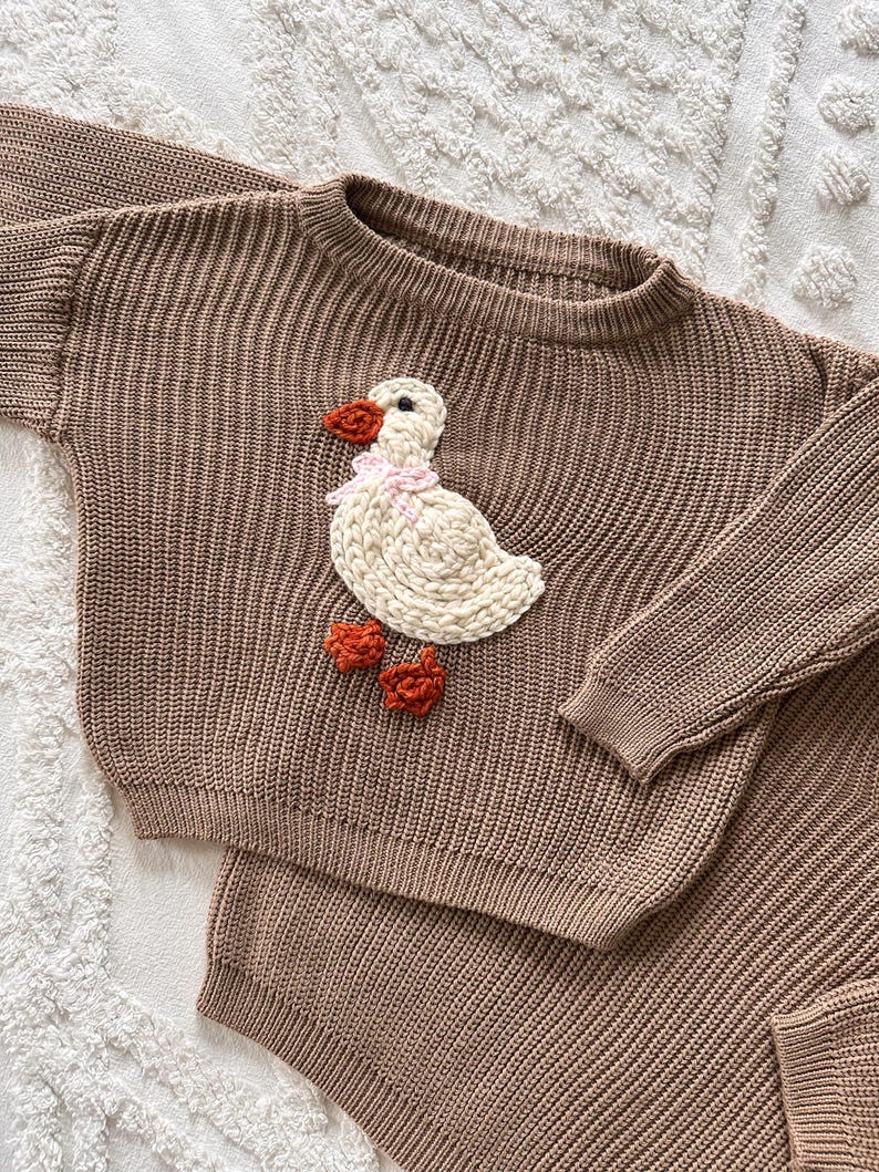 May include: A brown knit sweater and shorts set. The sweater features a white duck applique with an orange beak and feet, and a pink bow. The set is made of a ribbed knit fabric.