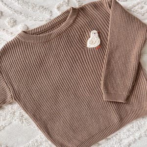 May include: A brown knit sweater with a crew neck and long sleeves. A white and orange embroidered bird is on the left chest. The sweater is made of a ribbed knit fabric.
