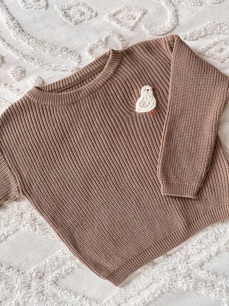 May include: A brown knit sweater with a crew neck and long sleeves. A white and orange embroidered bird is on the left chest. The sweater is made of a ribbed knit fabric.