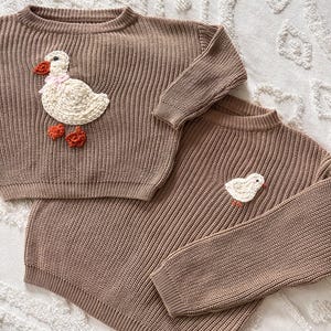 May include: Two brown knit sweaters. One sweater features a large white duck applique with a pink scarf and orange feet. The other sweater has a smaller duck applique. Both sweaters have ribbed texture.