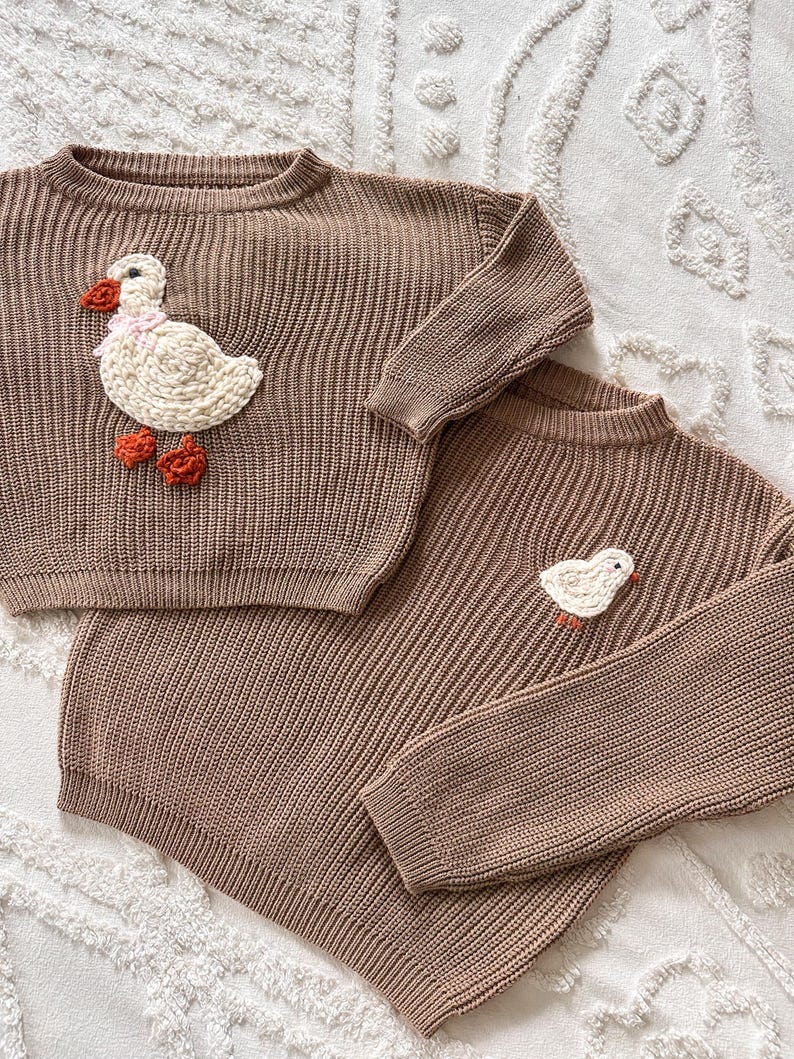May include: Two brown knit sweaters. One sweater features a large white duck applique with a pink scarf and orange feet. The other sweater has a smaller duck applique. Both sweaters have ribbed texture.
