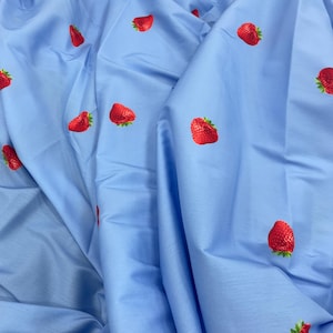 May include: Light blue fabric with a repeating pattern of red strawberries with green leaves. The fabric is draped, creating folds and shadows. The strawberries are evenly spaced across the material.