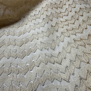 Puede incluir: Primer plano de una tela color crema con un patrón de chevrón repetido. La tela parece ser un encaje delicado o un tejido abierto, con una superficie ligeramente texturizada. El patrón crea una sensación de movimiento e interés visual.