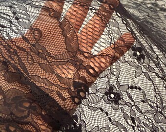 May include: Close-up of black lace fabric. The delicate material features intricate floral and geometric patterns. The lace is held up, showing the texture and transparency of the fabric. Ideal for apparel and craft projects.