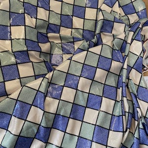 Peut inclure: Un tissu à motifs losanges en bleu, vert et crème, soulignés de noir. Le tissu est drapé, révélant des plis et une texture. Il présente un léger éclat, suggérant une matière soyeuse.