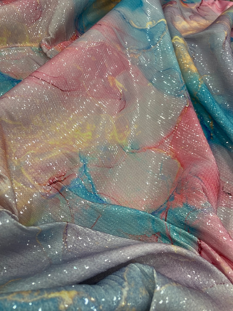 May include: A close-up of iridescent fabric with a marbled pattern in shades of pink, blue, and yellow. The fabric is covered in sequins that shimmer in the light.