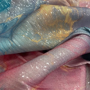 May include: A close-up of a soft, iridescent fabric with a textured, bubble-like surface. The fabric is folded and draped, revealing shades of blue, pink, and yellow.