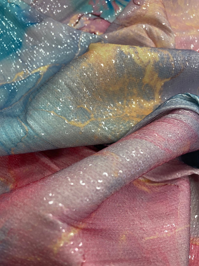 May include: A close-up of a soft, iridescent fabric with a textured, bubble-like surface. The fabric is folded and draped, revealing shades of blue, pink, and yellow.