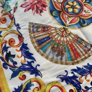 Può includere: Un tessuto bianco con un motivo floreale e geometrico colorato. Il design presenta un ventaglio blu e giallo, un fiore rosso e blu e un design geometrico giallo e blu.