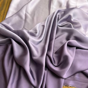 May include: Close-up of a draped fabric in shades of lavender. The fabric has a smooth, silky texture with soft folds and shadows. The colour transitions from a light lavender to a deeper, richer hue.