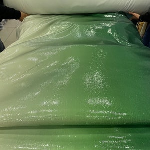 Puede incluir: Un rollo de tela verde iridiscente con un sutil efecto degradado. La tela está hecha de lentejuelas y tiene un acabado brillante.