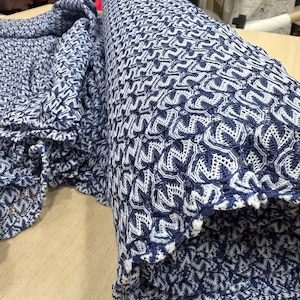May include: A roll of navy and white patterned fabric. The fabric has a repeating geometric design. The edge of the fabric has a scalloped trim. The fabric is on a wooden surface.