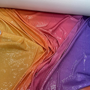 Peut inclure: Un rouleau de tissu avec un dégradé de couleurs allant du jaune à l'orange, au rose et au violet. Le tissu est recouvert de paillettes, créant un effet scintillant. Le bord du tissu a une bordure blanche.
