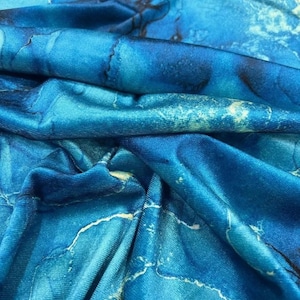 Op de afbeelding: Close-up van een stof met een marmerpatroon in blauwtinten. De stof heeft een vloeiende, gerimpelde textuur met witte en donkerblauwe aderingpatronen. Het algehele effect doet denken aan oceaangolven.