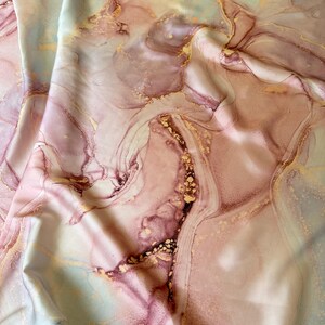 Puede incluir: Una tela con un diseño abstracto en tonos rosa, malva y dorado, con toques de azul pálido. La tela tiene una textura suave y sedosa, con un efecto marmolado. El diseño es fluido y orgánico.